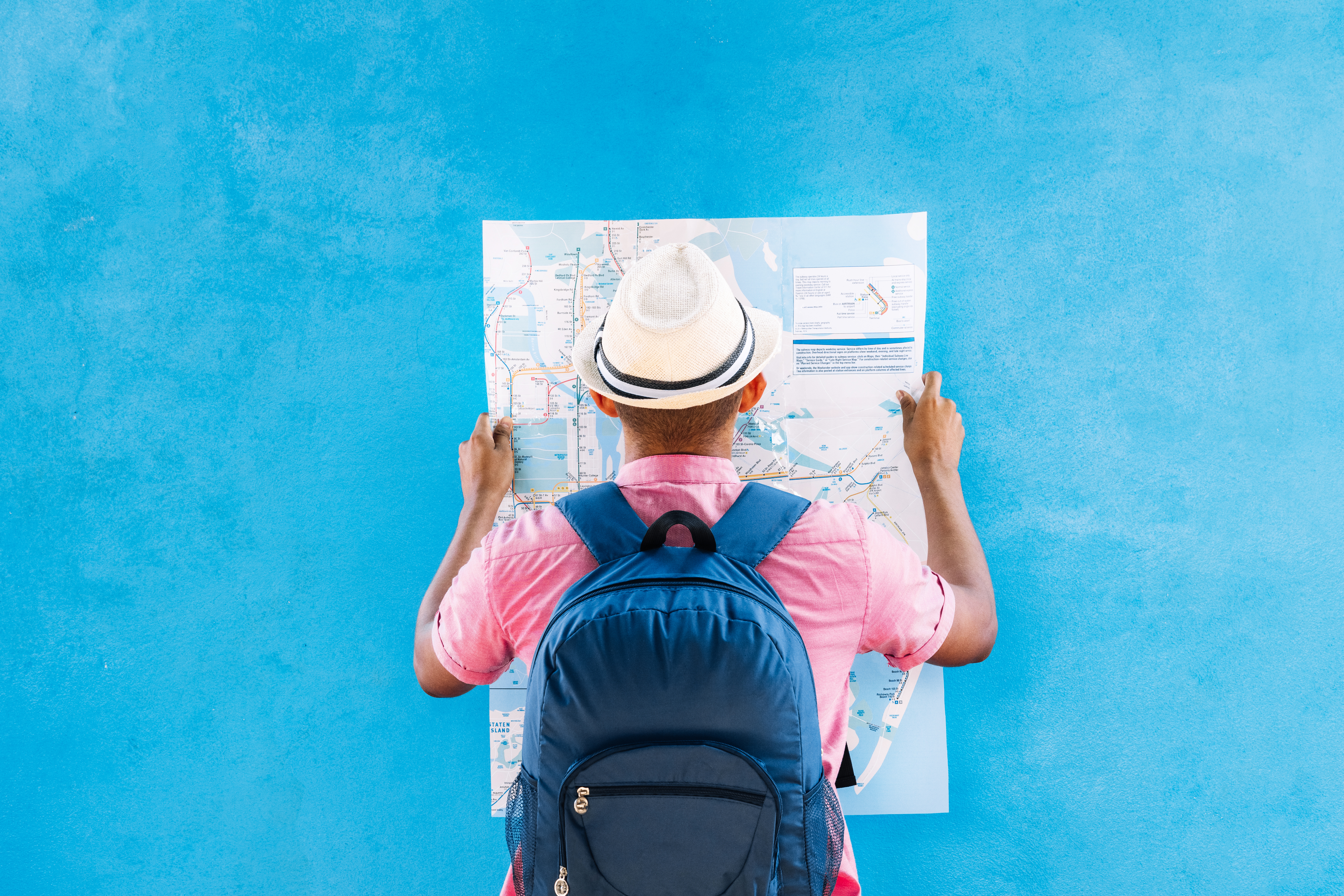 unrecognizable man with a sad travel hat and backpack, looking at a map, in front of a blue street wall. travel concept.